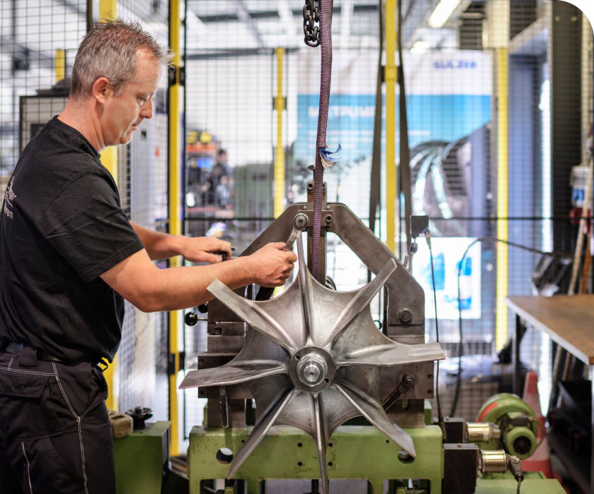 Ein Arbeiter in einem schwarzen T-Shirt bearbeitet ein großes metallisches Bauteil an einer Maschinenvorrichtung in einer Werkstatt.