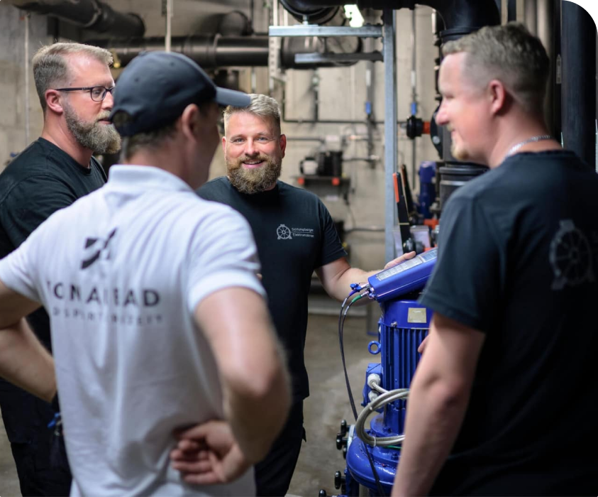 Vier Männer stehen in einer Werkstatt und konzentrieren sich auf eine blaue Industriemaschine. Einer lächelt und spricht, die anderen hören aufmerksam zu.